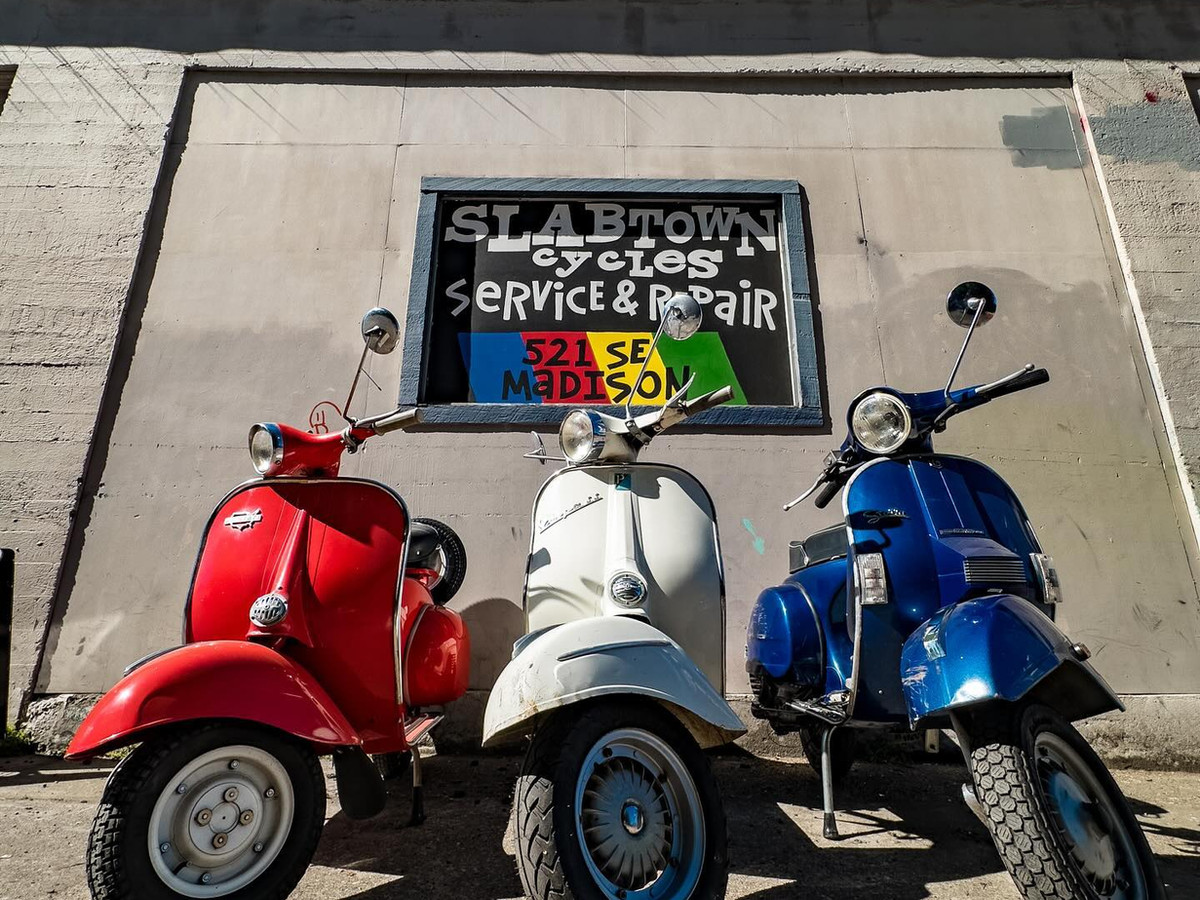 Three vintage Vespas — red, white, and blue — lined up in front of the hand-painted Slabtown shop sign